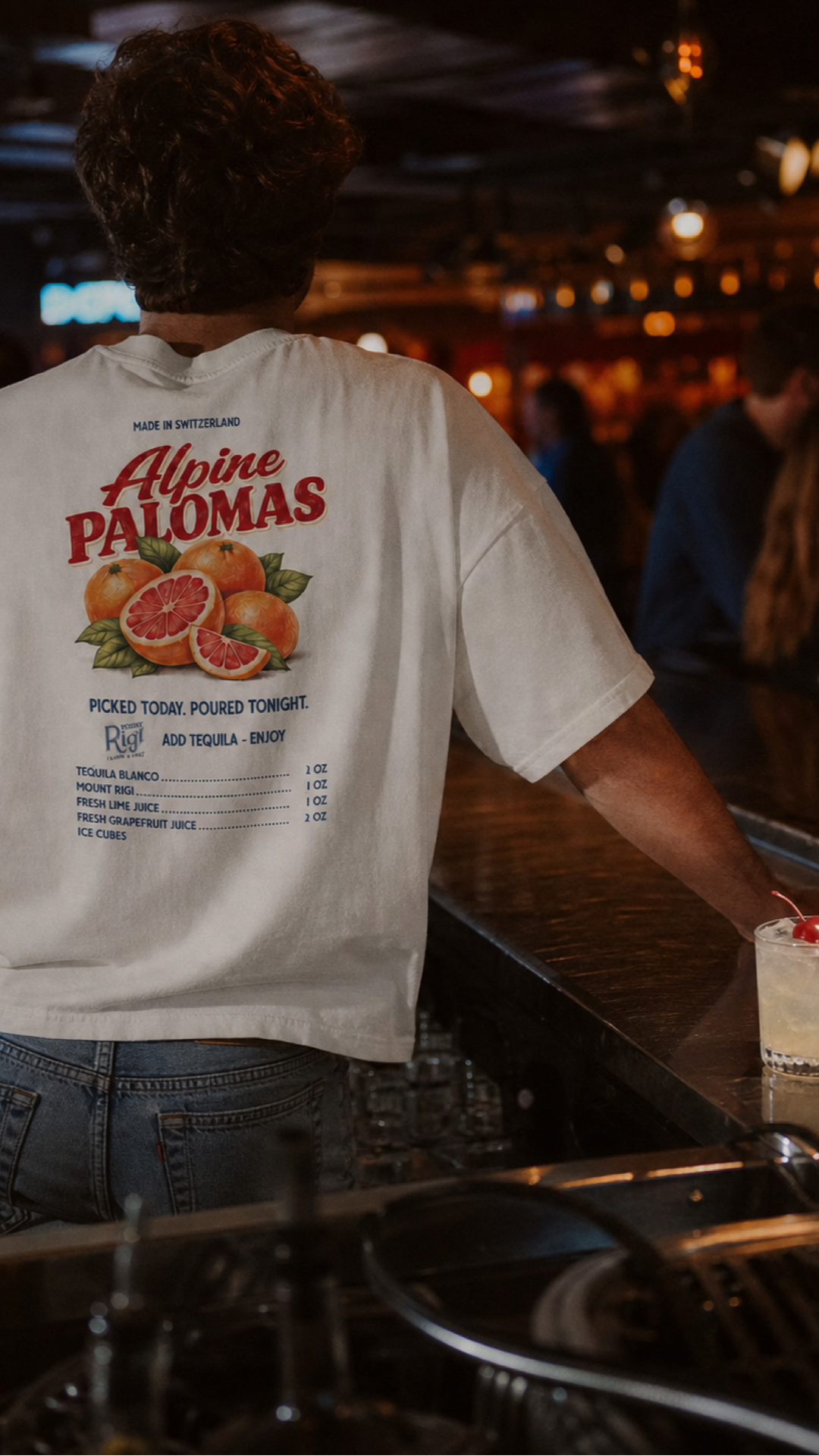 Man wearing the Alpine Palomas oversized T-shirt at a warmly lit bar with a cherry-garnished Paloma on the counter