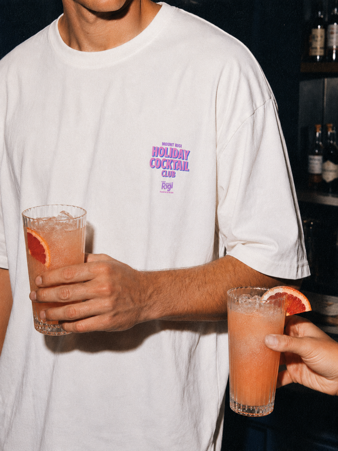 Man wearing a Mount Rigi Holiday Cocktail Club T-shirt holding a grapefruit cocktail, with a second cocktail in the foreground.